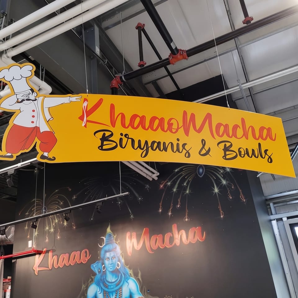 Hanging yellow Khana Machna Biryani & Bowls sign featuring a cartoon chef inside an industrial restaurant.