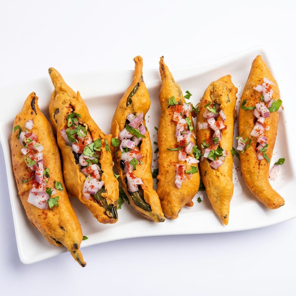 Six golden-fried chili pakoras, garnished with chopped red onions, cilantro, and red spice, on a white plate.