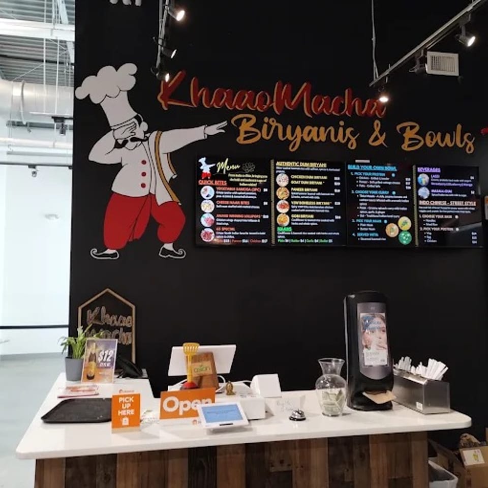Restaurant counter featuring a chef mural, biryanis and bowls menu boards, self-service station, and open sign.